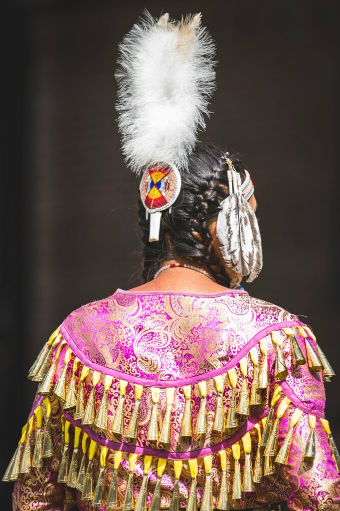 Photo of the back of a women in a pink jingle dress. She has dark brown hair with a white feather in it.